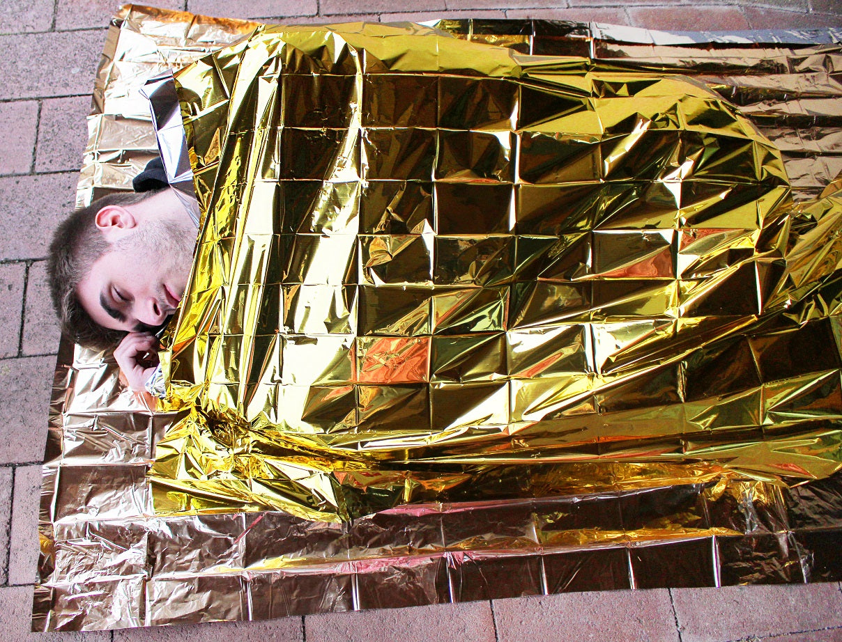 Das Bild zeigt eine Rettungsdecke, die aus zwei Seiten besteht: eine goldene und eine silberne. Die Rettungsfolie hat eine Größe von 160 x 210 cm und ist zusammengefaltet. Der glänzende, reflektierende Stoff ist leicht erkennbar und scheint in der Mitte leicht zerknittert zu sein. Im Hintergrund ist ein neutraler, verschwommener Untergrund zu sehen, der dem Produkt Raum gibt und den Fokus auf die Rettungsdecke lenkt. Diese Decke ist ideal für Notfälle und hilft bei der Thermoregulation.