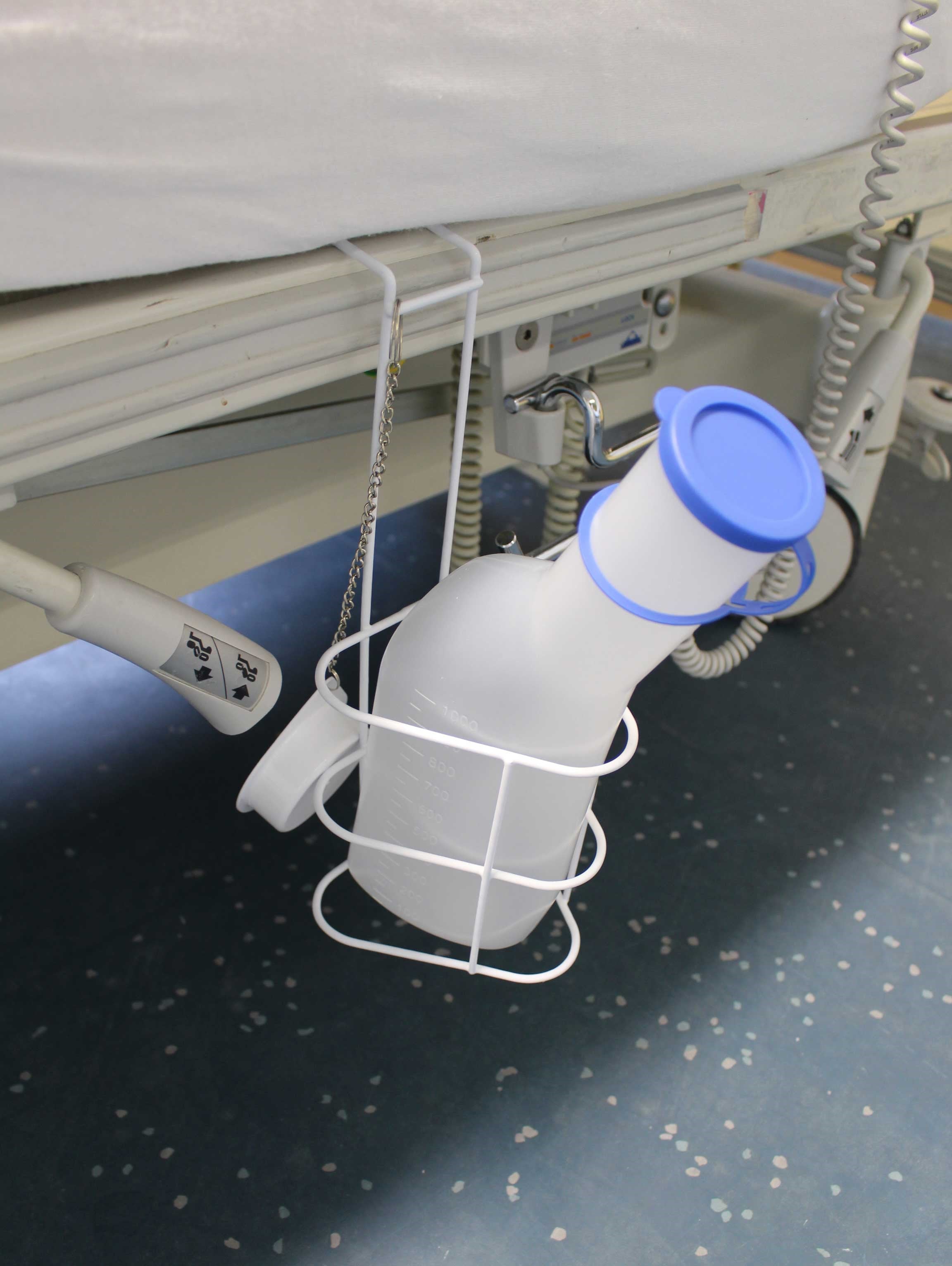 Urinflasche aus milchigem Polypropylen mit einem Fassungsvermögen von 1000 ml, speziell für Männer. Die Flasche hat eine konische Form mit einer breiten Öffnung für einfache Handhabung. Ein abnehmbarer Betthalter ist dabei, der die Flasche sicher hält. Im Bild sind auch die integrierte Reinigung Bürste und ein handlicher, benutzerfreundlicher Deckel zu sehen. Die gesamte Anordnung ist auf einem weißen Hintergrund, was die Produktdetails hervorhebt und die Benutzerfreundlichkeit unterstreicht.