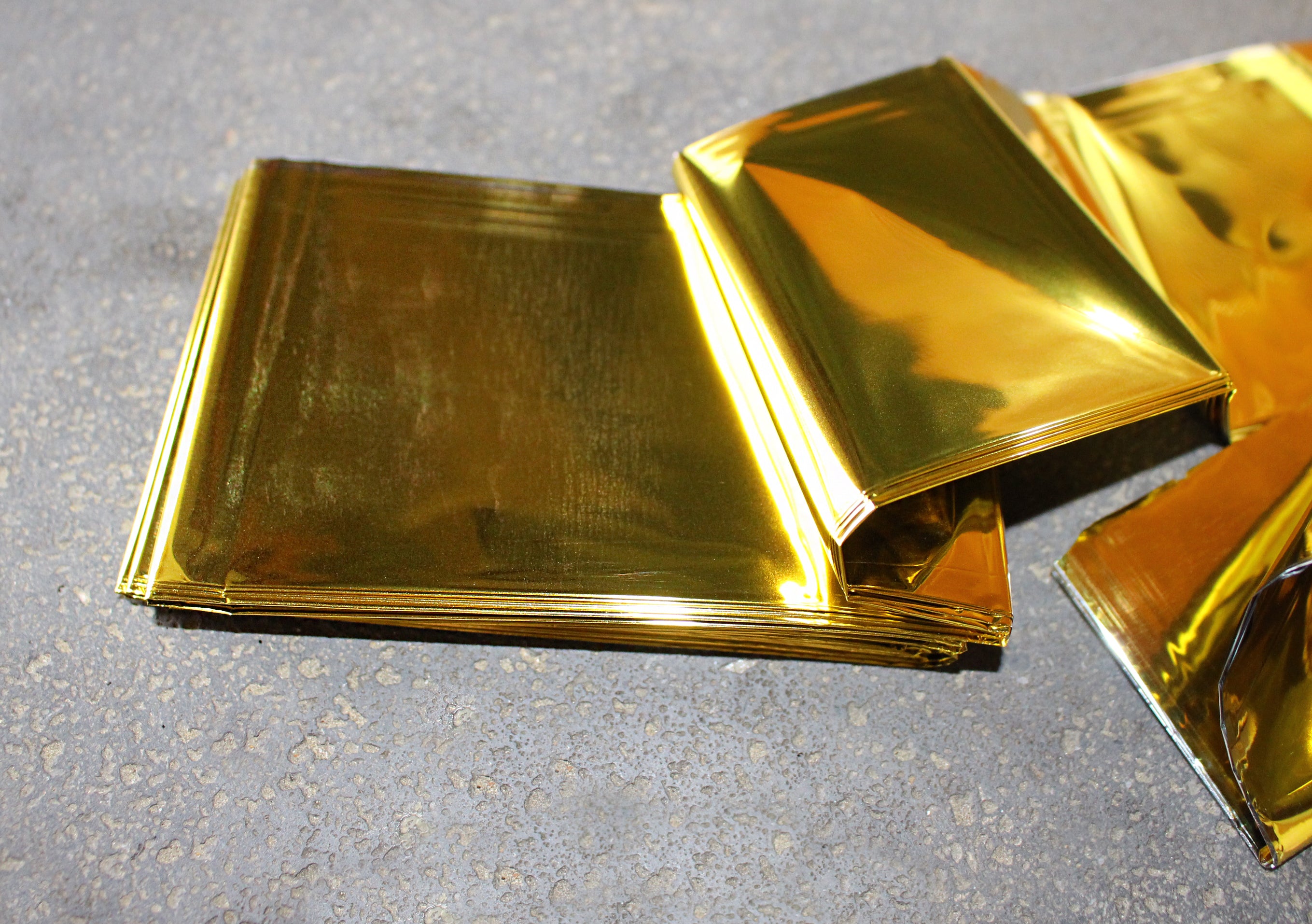 Bild eines Satzes von zwei Noba-Rettungsdecken in Gold und Silber, jeweils in der Größe 1,6 m x 2,1 m. Die Decken sind sorgfältig gefaltet und liegen übereinander auf einem neutralen Hintergrund. Die goldene Decke ist auf der Oberseite sichtbar, während die silberne Decke darunter liegt. Beide Decken strahlen eine glänzende Oberfläche aus, die das Licht reflektiert.