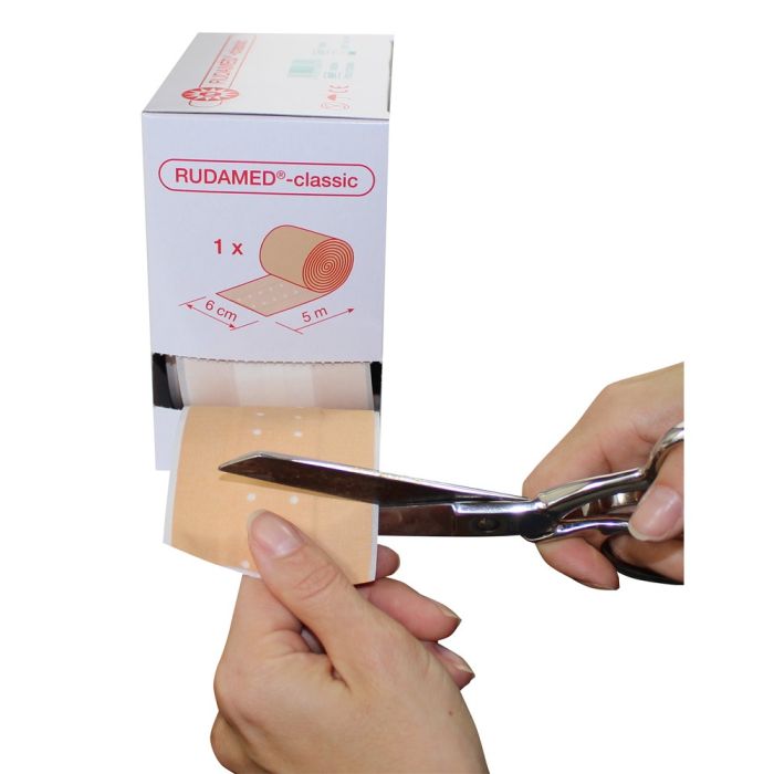 Ein Noba Rudamed Classic Wundpflaster ist auf einem weißen Hintergrund abgebildet. Das Produkt zeigt eine Rolle von Wundschnellverband, die etwa 5 Meter lang ist. Die Verpackung ist klar und farbenfroh gestaltet, mit dem Markennamen "Noba" und dem Produktnamen "Rudamed" prominent in leuchtendem Blau und Gelb. Auf der Vorderseite sind zudem Hinweise zur Anwendung und ein Symbol für Wundschutz abgebildet. Die Rolle selbst ist unentfernt und die Kante des Pflasters ist sichtbar.