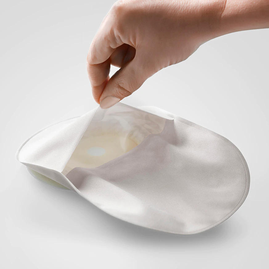 Sensura Mio Kolostomiebeutel in lichtgrauer Farbe, Größe 15-45 mm, maximal 510 ml Fassungsvermögen. Der Beutel hat eine abgerundete Form und eine matte Oberfläche. Auf der oberen Kante sind transparente Bereiche für eine einfache Befestigung an Hautschutzplatten zu sehen. Die Rückseite des Beutels zeigt ein flexibles Design, das sich an die Körperform anpasst. Der Beutel ist für eine diskrete Anwendung konzipiert und genügt hohen hygienischen Standards. Ideal für Menschen mit Kolostomie.