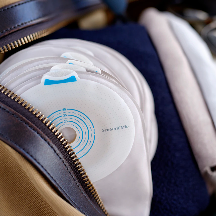 Ein Lichtgrauer Sensura Mio Kolostomiebeutel mit einem Plan von 30 mm Durchmesser ist abgebildet. Der Beutel hat eine glatte, matte Oberfläche und eine ergonomische Form, die sich der Körperkontur anpasst. Die obere Kante des Beutels zeigt eine selbstklebende Oberfläche für die sichere Anbringung an der Haut. Der Beutel ist leicht transparent und zeigt eine subtile Textur. Auf der Rückseite sind Informationen und ein Herstellerlogo zu erkennen.