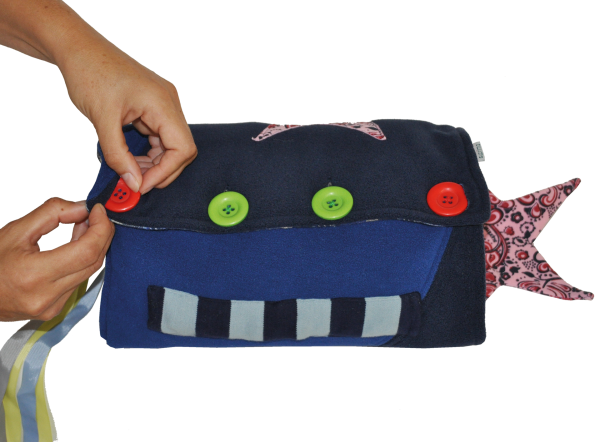 Bild eines demenzfreundlichen Handmuffs von Suprima, der auf einem hellen Hintergrund präsentiert wird. Der Muff ist in sanften Farben gehalten, überwiegend in Grau, mit bunten Elementen wie Stoffapplikationen und verschiedenen Texturen. Er ist mit kleinen Spielzeugen und Knöpfen ausgestattet, die die taktile Stimulation fördern. Der Handmuff ist zweigeteilt, ideal für die Hände von Senioren.