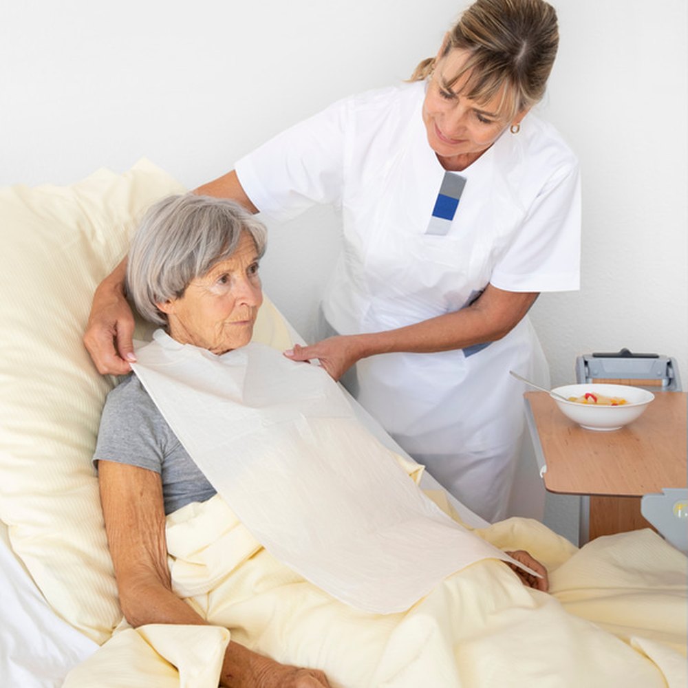 Pflegekraft legt einer älteren Frau im Bett einen Einweg-Esslatz an.